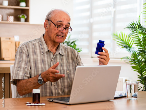 Senior man having an online health consultation on laptop at his home, discussing medication, capsule dosage, illness prevention and medical support.