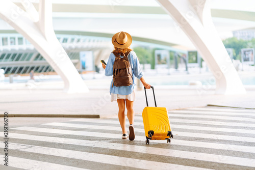 Woman wearing hat and carrying backpack stands with yellow suitcase in a modern city on a sunny day. Tourist with phone enjoys a trip at sunset. Concept for tourism, recreation, and blogging.