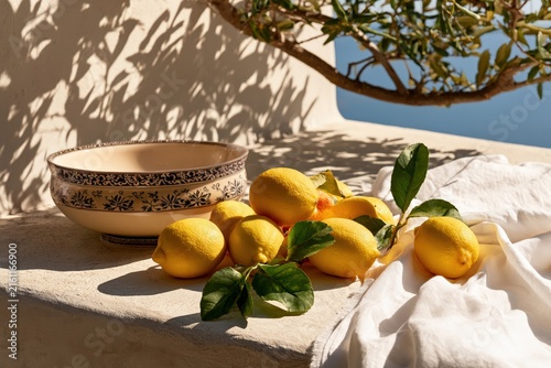 sunlit mediterranean still life with fresh lemons, green leaves, decorative ceramic bowl and white linen on a rustic terrace with olive tree shadows