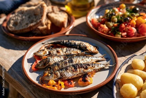 traditional Portuguese grilled sardines with roasted peppers, boiled potatoes, rustic bread and fresh tomato olive salad served on a sunlit wooden table