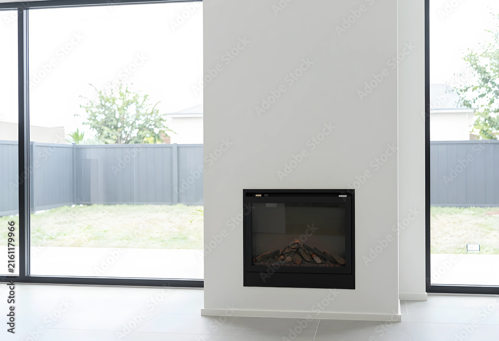 Fototapeta premium Modern home interior with minimalist design, built-in electric fireplace and large glass windows overlooking a backyard
