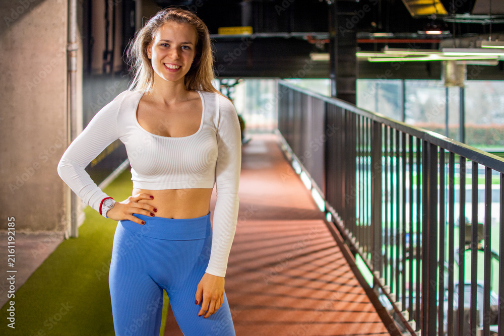 Fototapeta premium Smiling fit woman standing near balcony railing posing confidently in modern gym.