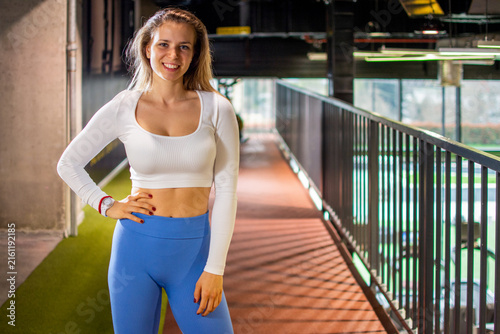 Smiling fit woman standing near balcony railing posing confidently in modern gym.