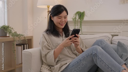 Asian woman using smartphone sofa bright living room home, smiling during casual mobile communication, relaxing indoors with houseplant, soft daylight, neutral decor, comfortable furnishings and calm