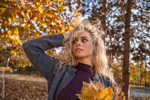 Closeup shot of natural looking beautiful young blonde with curly hair holding dry autumn leaves at sunset 