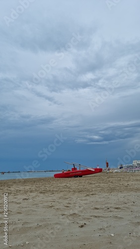 Red rescue boat resting on a wide sandy beach beneath a dramatic overcast sky, with the calm sea visible on the horizon.