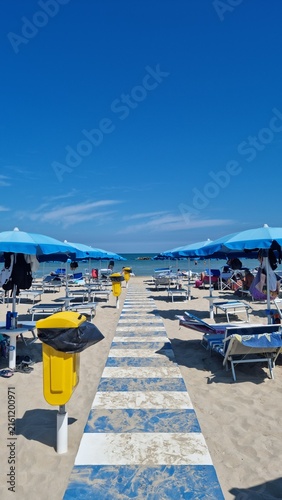 Sunny beach resort with blue umbrellas, sun loungers and a tiled walkway leading toward the sea under a clear blue sky.