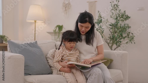 Mother and daughter reading colorful vdo book together on cozy living room sofa, sharing tender family moment with child and woman at home in warm peaceful interior