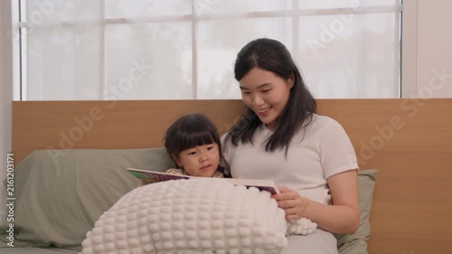 Asian mother and daughter reading storybook together on bed in bright bedroom, warm family moment with child and woman sharing gentle smiles beneath textured blanket at bedtime