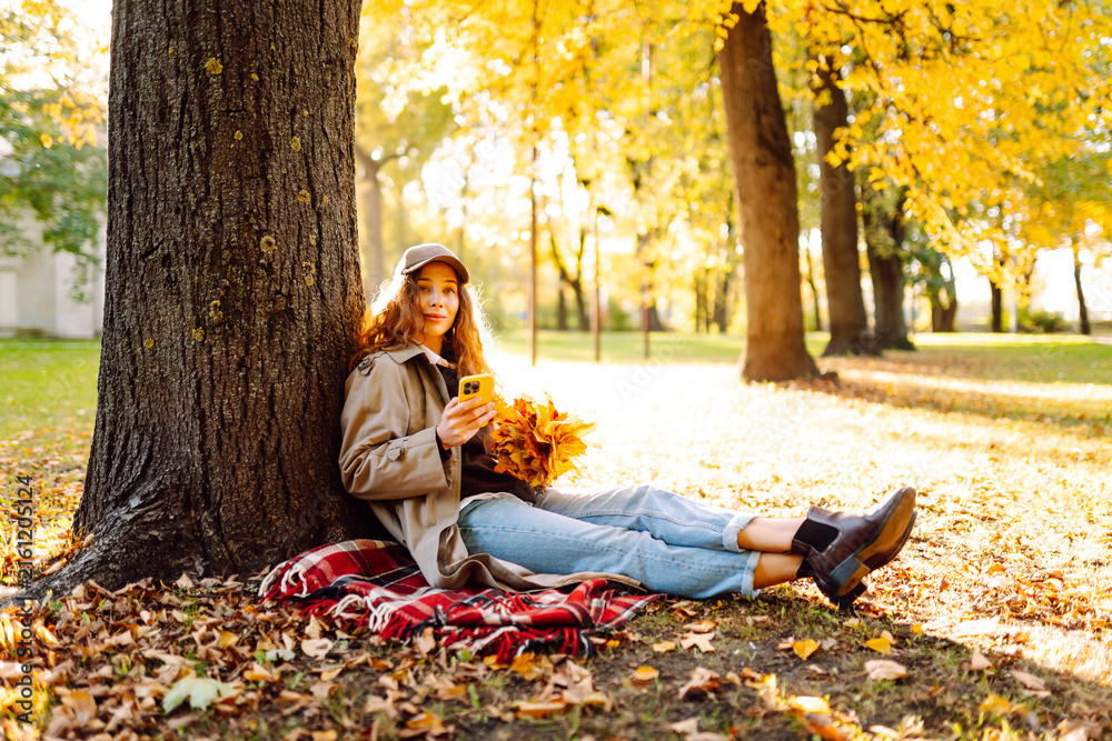 Fototapeta premium Woman sits on blanket near tree with phone and bouquet of bright yellow leaves. Woman relaxes in autumn park at sunset with smartphone. Concept of seasonality, relaxation. Lifestyle.