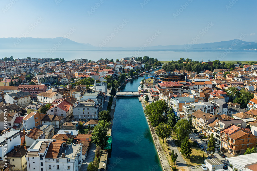 Fototapeta premium Aerial drone view of Struga in North Macedonia with Black Drin River flowing into crystal clear Lake Ohrid, boats, sunny summer landscape, waterfront buildings and scenic city panorama