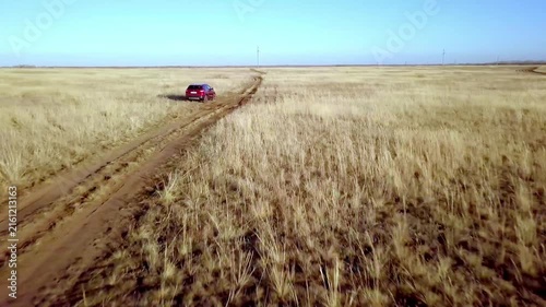 Aerial view of red SUV car driving on dirt road through dry grassy autumn field
