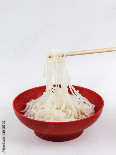 Chopsticks lifting translucent shirataki or konjac noodles from a vibrant red bowl against a clean white background.