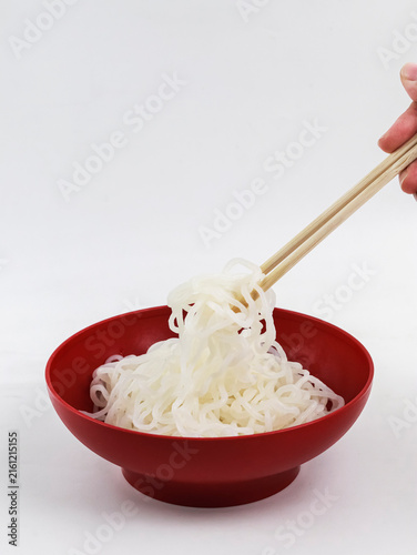 A hand holding wooden chopsticks lifts translucent shirataki or konjac noodles from a vibrant red bowl against a clean white background.