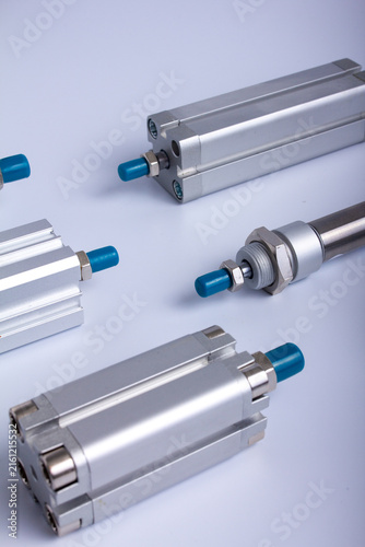 Top view close up of various industrial pneumatic air cylinders and aluminum actuators with blue caps laid out on a white surface for automated machinery systems.