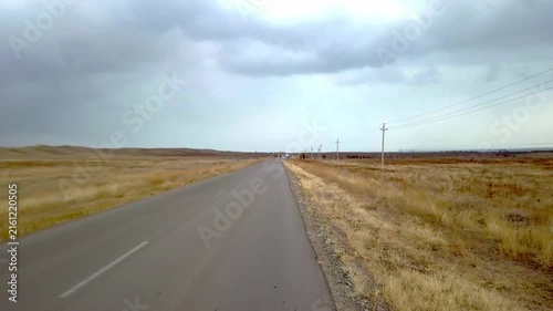 Asphalt road stretching through dry grassy steppe under dramatic overcast cloudy sky, driving perspective