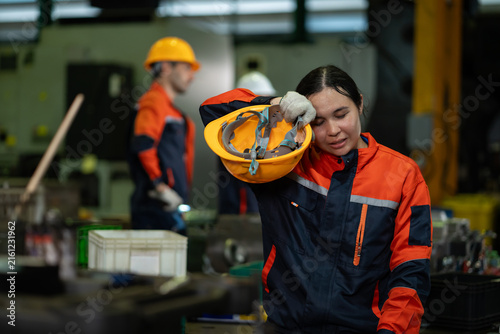 Exhausted female factory worker resting with removed safety helmet, showing fatigue and tiredness at an industrial workplace,  overwork, stress and labor exhaustion