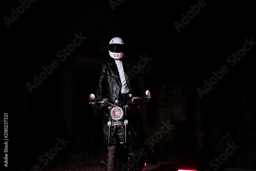 Tired male motorcyclist in white helmet and leather jacket resting on ground next to motorcycle wheel at dark night. Road trip fatigue and night travel concept