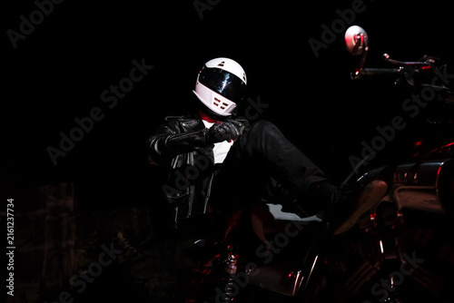 Tired male motorcyclist in white helmet and leather jacket resting on ground next to motorcycle wheel at dark night. Road trip fatigue and night travel concept