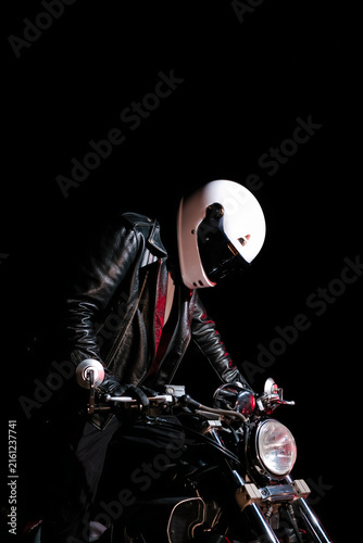 Tired male motorcyclist in white helmet and leather jacket resting on ground next to motorcycle wheel at dark night. Road trip fatigue and night travel concept