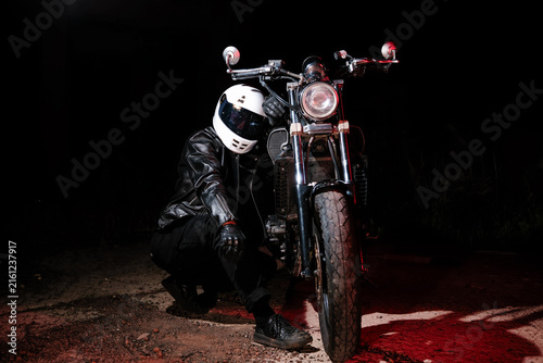 Tired male motorcyclist in white helmet and leather jacket resting on ground next to motorcycle wheel at dark night. Road trip fatigue and night travel concept