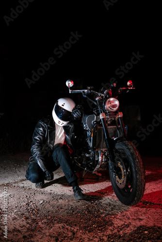 Low key portrait of motorcyclist in white helmet and black leather jacket sitting on motorcycle in dark night. Night ride, dramatic lighting and biker lifestyle concept