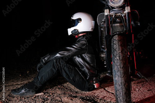 Low key portrait of motorcyclist in white helmet and black leather jacket sitting on motorcycle in dark night. Night ride, dramatic lighting and biker lifestyle concept
