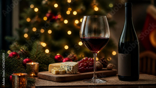 Glass of red wine and a cheese board with grapes on a rustic table with a blurred christmas tree background and warm festive lights