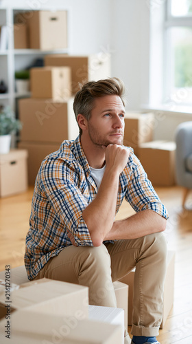 Thoughtful young Caucasian man pondering while surrounded by moving boxes.