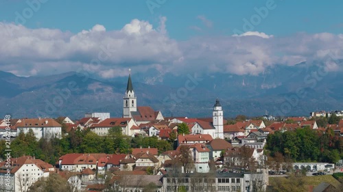 Aerial view shows Kranj, Slovenia with its medieval old town, church towers, and surrounding landscape. Clear blue sky highlights the scene.