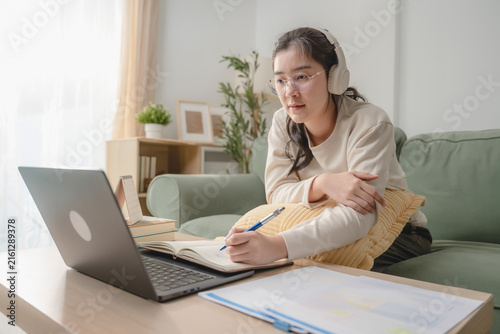 Asian woman wearing headphones writing in notebook while studying online course on laptop at home, concept of e-learning and distance education