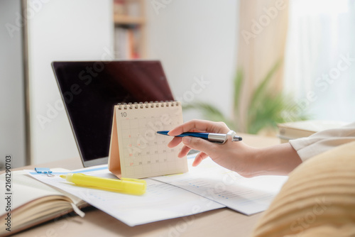 Close up hand holding pen pointing at desk calendar with laptop and papers on table, concept of project planning, schedule management, time management at home and deadline tracking.