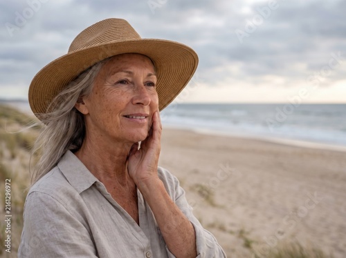 Elderly woman wearing straw hat and linen shirt standing on sandy beach coastline overlooking ocean