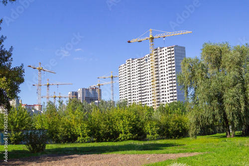 White building and construction cranes among trees against blue sky