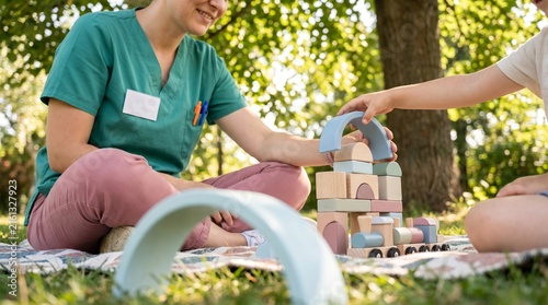 Pediatric nurse playing with child outdoors, healthcare worker using wooden blocks for therapy, medical professional in green scrubs