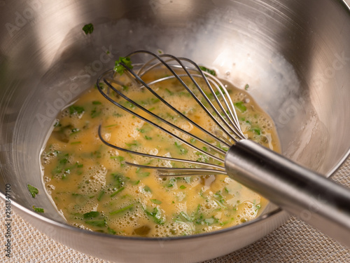 A whisk blends golden beaten eggs with flecks of fresh parsley in a shiny stainless steel bowl. The close angle shows the frothy texture forming during meal preparation.