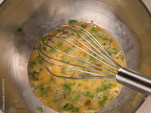 A metal whisk stirs beaten eggs mixed with chopped fresh parsley inside a stainless steel mixing bowl. Small bubbles form on the surface as the egg mixture is prepared for cooking.