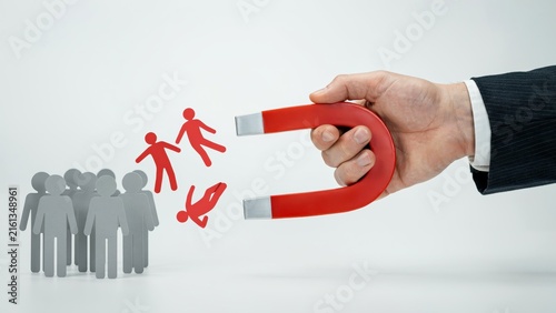 A hand holding a red horseshoe magnet attracting red paper people icons away from a grey group isolated on white background. Lead generation business concept.
