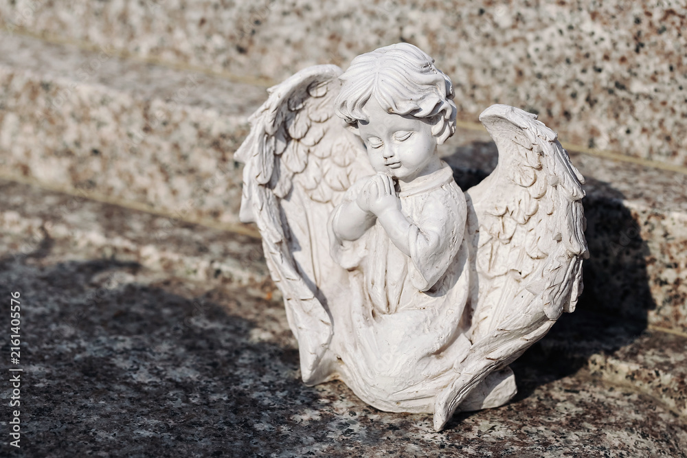 Fototapeta premium White praying angel statue rests outdoors. Decorative wings surround peaceful child figure. Religious ornament stands on stone surface. Memorial sculpture forms spiritual background.