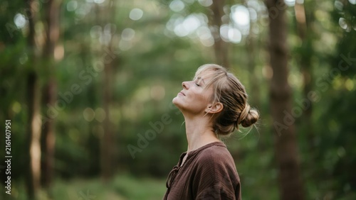 A young woman standing in a forest with her eyes closed, taking a deep breath and enjoying the peaceful nature and fresh air outdoors