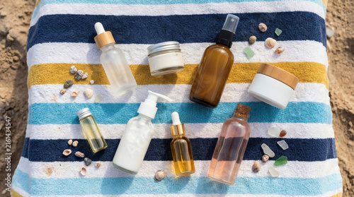Various skincare and cosmetic bottles, jars, and vials are arranged on a colorful striped beach towel on the sand, accessorized with seashells.