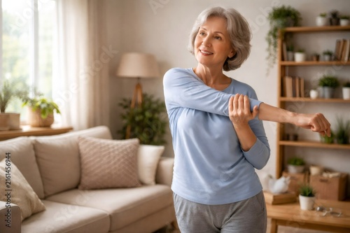 Senior woman with gray hair smiling, stretching arm across her chest, practicing daily routine for healthy body and active lifestyle in bright home interior