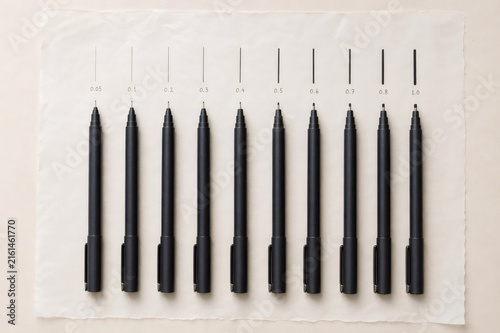Row of black fineliner pens displaying various tip sizes from 0.05 to 1.0, each pen aligning with its corresponding line weight example on crumpled paper