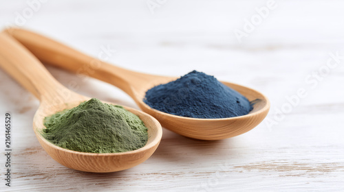 Wooden spoons filled with green spirulina and blue spirulina powders placed on a light wooden surface, showcasing vibrant colors and textures of the natural ingredients