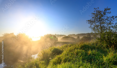 Misty dawn over river with sun breaking through trees and grass
