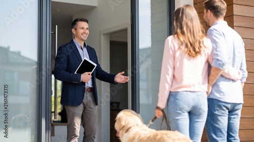 A real estate agent stands at the doorway, smiling and greeting two clients who are visiting for a home showing. The setting is bright and inviting, showcasing an interior