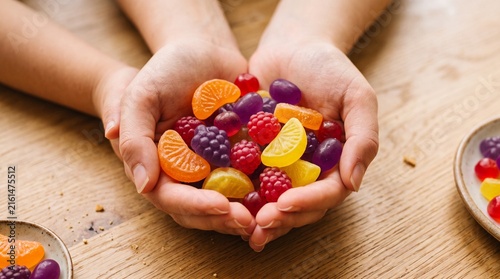 Two hands holding colorful gummy fruit candies, bright red, yellow, orange, purple pieces shaped like slices and berries, wooden surface with bowl and patterned cloth background.