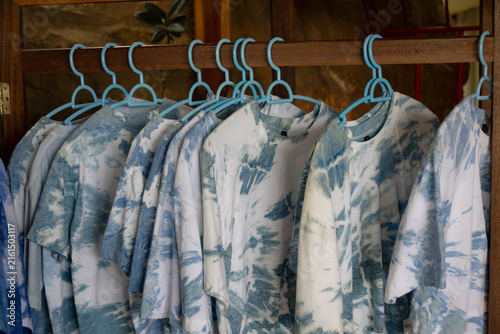 close-up shot of tie-dye t-shirts hanging on light blue plastic hangers. The shirts feature a soft, pastel tie-dye pattern.