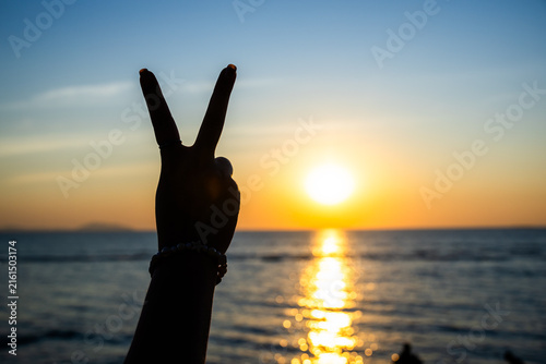 silhouetted hand making a peace sign against a glowing sunset over the ocean, with golden light