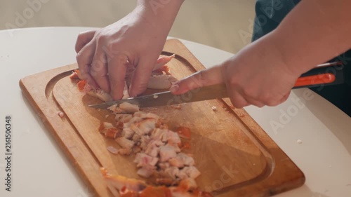 Close-up three-quarter top view shows human hands chopping cooked seafood into small pieces on wooden cutting board in bright home kitchen, with knife cutting and left hand holding ingredients